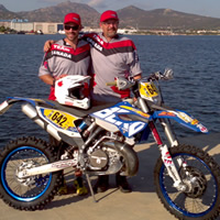 Two men standing in front  of a bay of water with their dirt bike. 