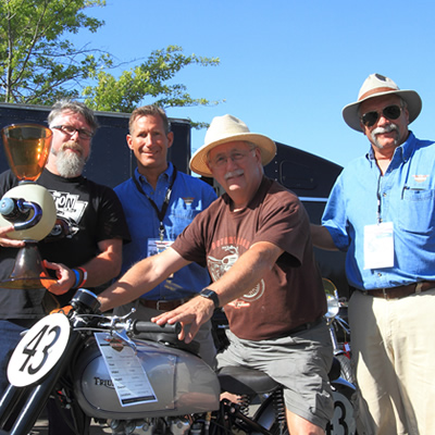 Picture of motorcycle enthusiasts, (one sitting on a motorcycle) and man holding trophy. 