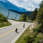 Three motorbikers drive down a sunny road with Kootenay Lake in the background.