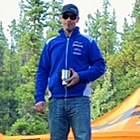 Photo of three men standing on a log podium with drinks in their hand and orange tents in the background. 