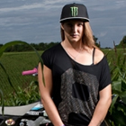 Photo of a girl with long blond hair standing in front of a dirt bike in a corn field. 