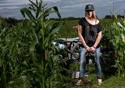 Photo of a girl with long blond hair standing in front of a dirt bike in a corn field. 