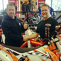 Two men shaking hands behind a KTM dirt bike. 