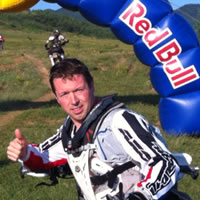 A brown haired man putting the thumbs up while sitting on his bike in front of Red Bull sign. 