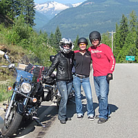 Three motorcycle riders standing by a Harley Davidson on the side of the road. 