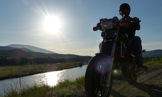 A silhouette of a guy on a motorcycle parked on a hill overlooking a river. 