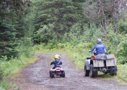 Man and a boy, each on an ATV, riding on a trail into the forest