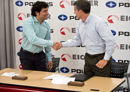 Photo of a man in a light blue long sleeve shirt shaking hands with another man who is in a white shirt. They are standing behind a desk. 