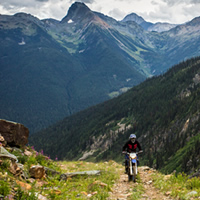 A man on a dirt bike riding up a rocky trail with the mountains in the background. 