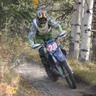 Photo of a guy coming around a corner in the trees on a dirt bike. 