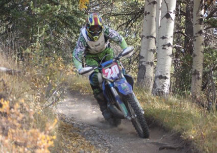 Photo of a guy coming around a corner in the trees on a dirt bike. 