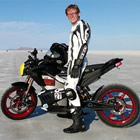 Photo of a young man dressed in white and black riding gear standing next to a black, red and white street bike. 